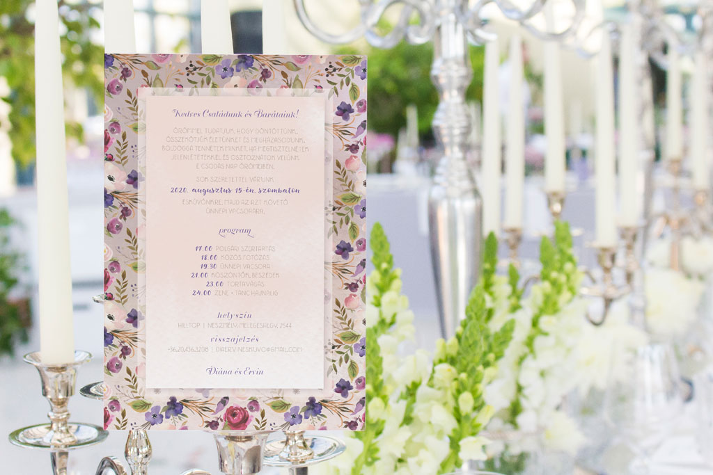 Levendula púder virágos esküvői meghívó élénk ciklámen és lila virágokkal, kevés zöld levéllel és halvány drapp, kézzel festett alnyomat grafikával.