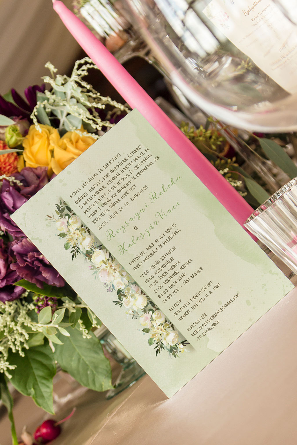 Fehér virágos esküvői meghívó halvány krém színű és törtfehér virágokkal, természetes zöld levelekkel és halvány zöld, vízfestékes alnyomat grafikával.