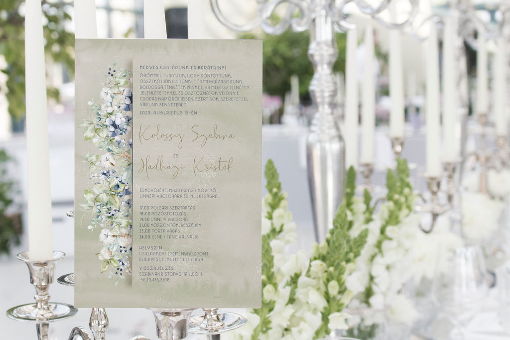 Téli virágos esküvői meghívó halvány szürkéskék apró virágokkal. Lila bogyós növénnyel és zöld levéllel. Zöldes szürke vízfestékes alnyomat grafikával.